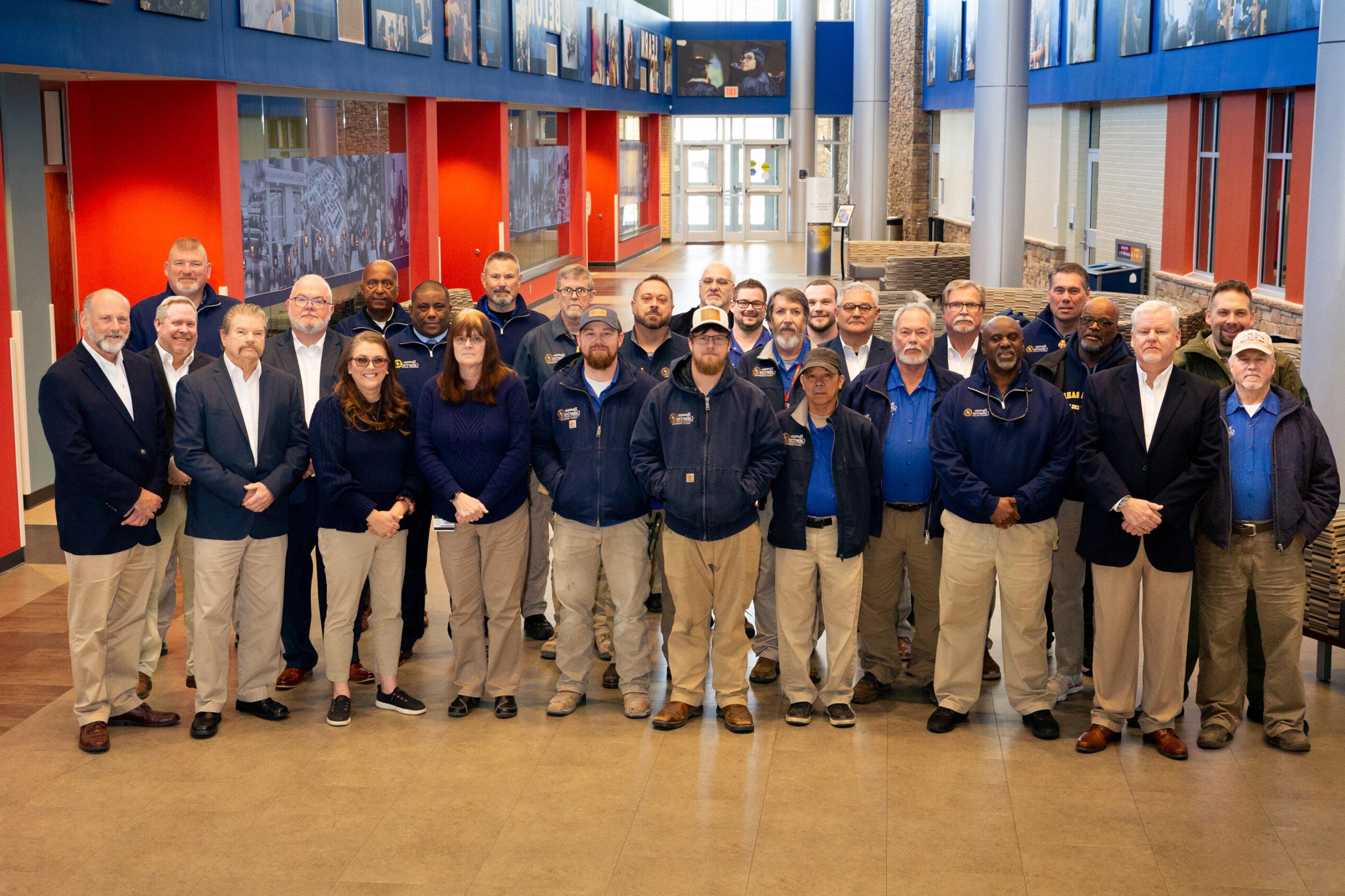 A group of 28 people in blue jackets or formal attire standing indoors with red walls and photo displays.