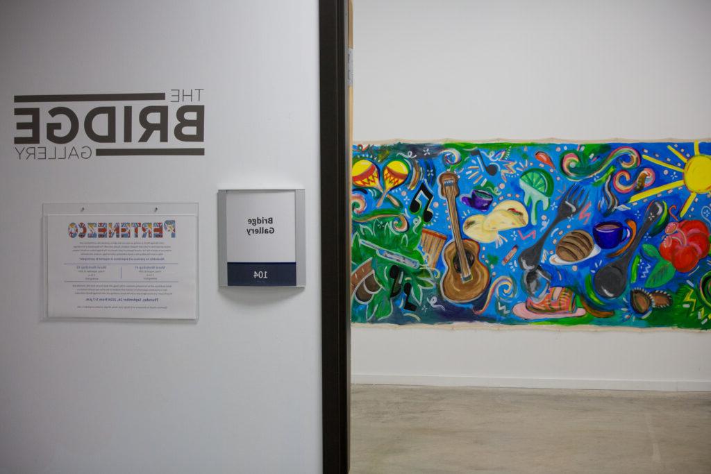 An open door leading into the Bridge Gallery (Room 104), with a vibrant and colorful mural visible inside. The mural features a mix of musical instruments, food items, and natural elements painted in a bold, dynamic style. On the wall outside the gallery, signage displays 'The Bridge Gallery' and a poster for the 'Pertenezco' workshop series.