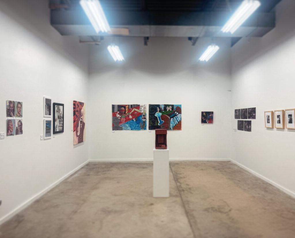 An interior view of the Bridge Gallery with bright fluorescent lights illuminating the space. The walls are adorned with an eclectic mix of artworks, including paintings, sketches, and photographs. A central pedestal displays a three-dimensional sculpture, while the concrete floor adds a modern, minimalist aesthetic to the gallery environment.