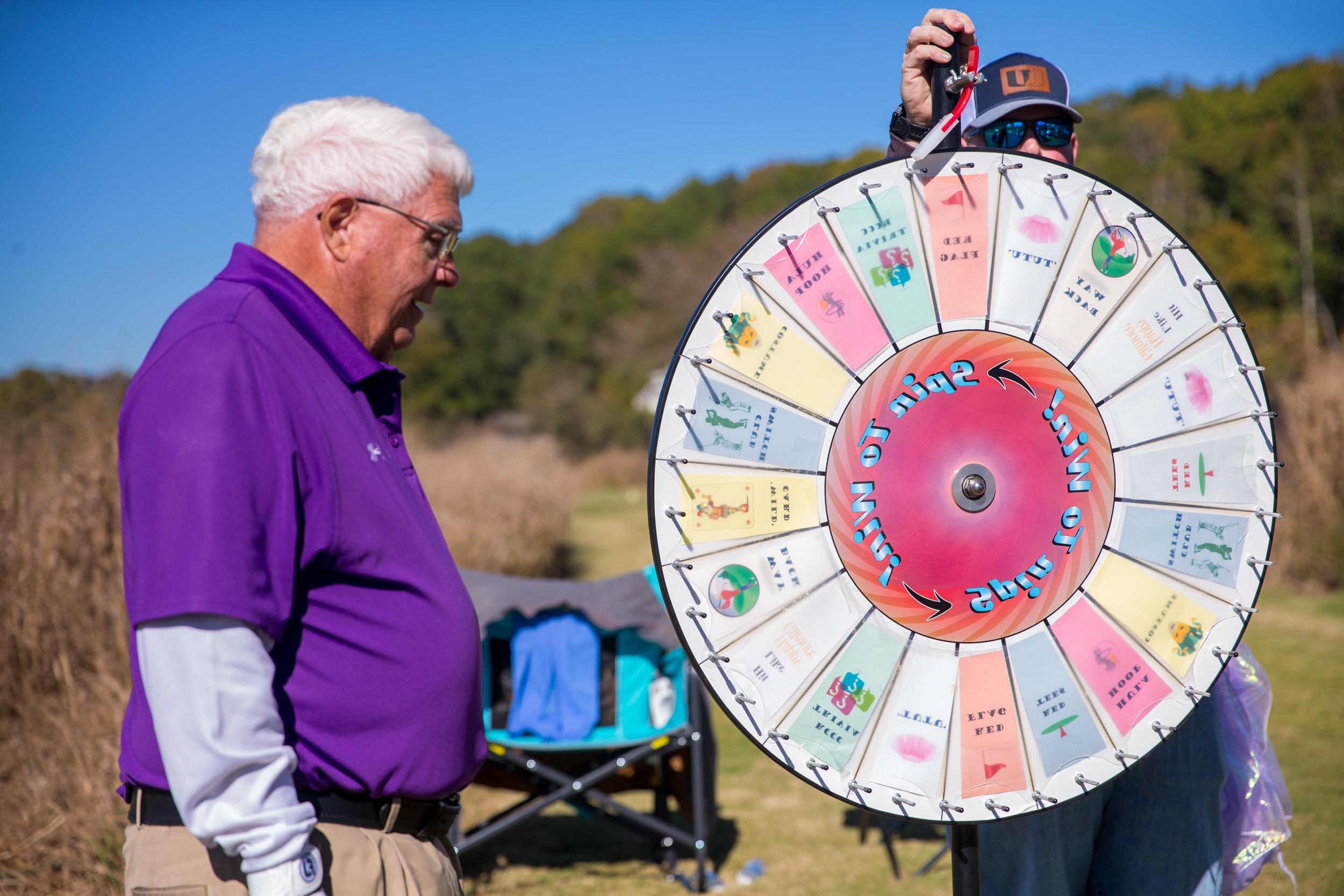 a man wearing a purple polo shirt standing outdoors beside a colorful spinning prize wheel. The wheel is large, with multiple triangular sections containing various labels and images. Another person, partially visible, is holding a handle to spin the wheel.