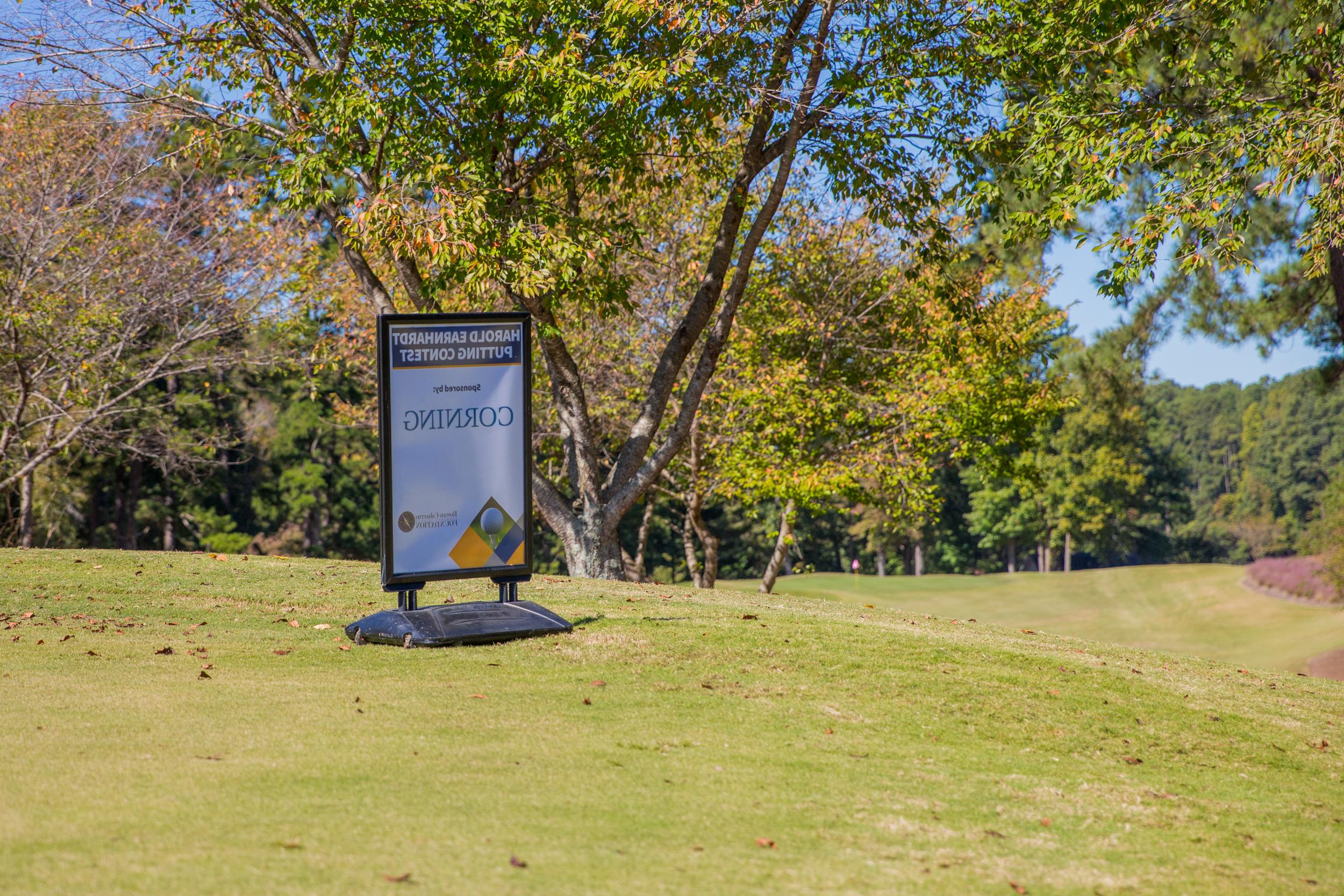 A sign on a grassy golf course surrounded by trees under a clear blue sky. Transcribed Text: HAROLD EARNHARDT PUTTING CONTEST Sponsored by: CORNING