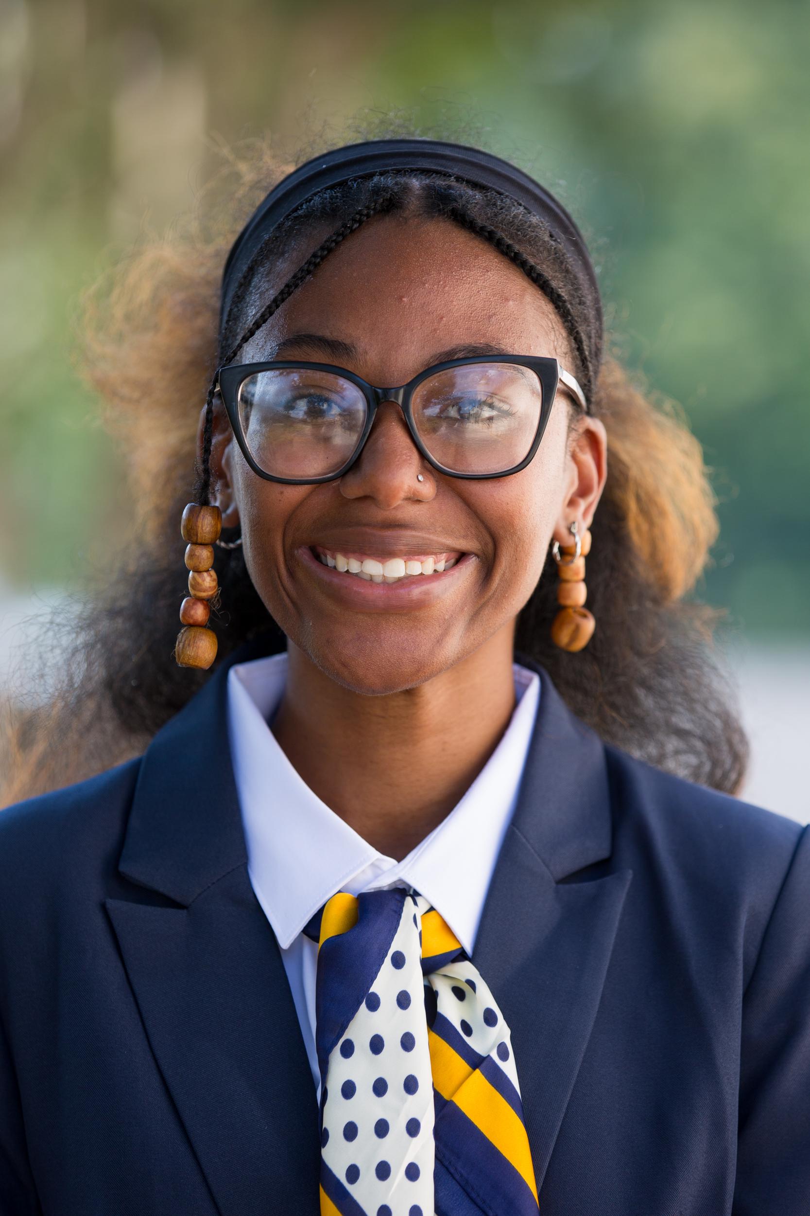 Destiny McPherson, wearing big black cat-eye glasses, has her two-tone brown hair pulled back with a black headband and a small braid pulled back on each side. She is dressed in a navy blazer, a white shirt, a navy and gold scarf, and hanging wooden earrings, completed by a smile.