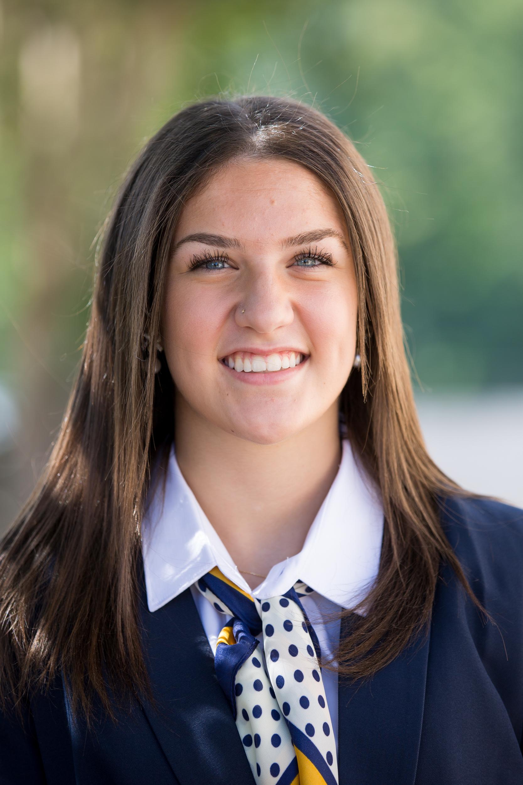 Jennifer Jellen<br />
 has a big smile with long, straight brown hair, wearing a professional dark blue jacket over a white shirt. She has a yellow, white, and blue polka-dot patterned scarf around her neck. she has a small stud in her nose and dangling pearl and bow earrings.