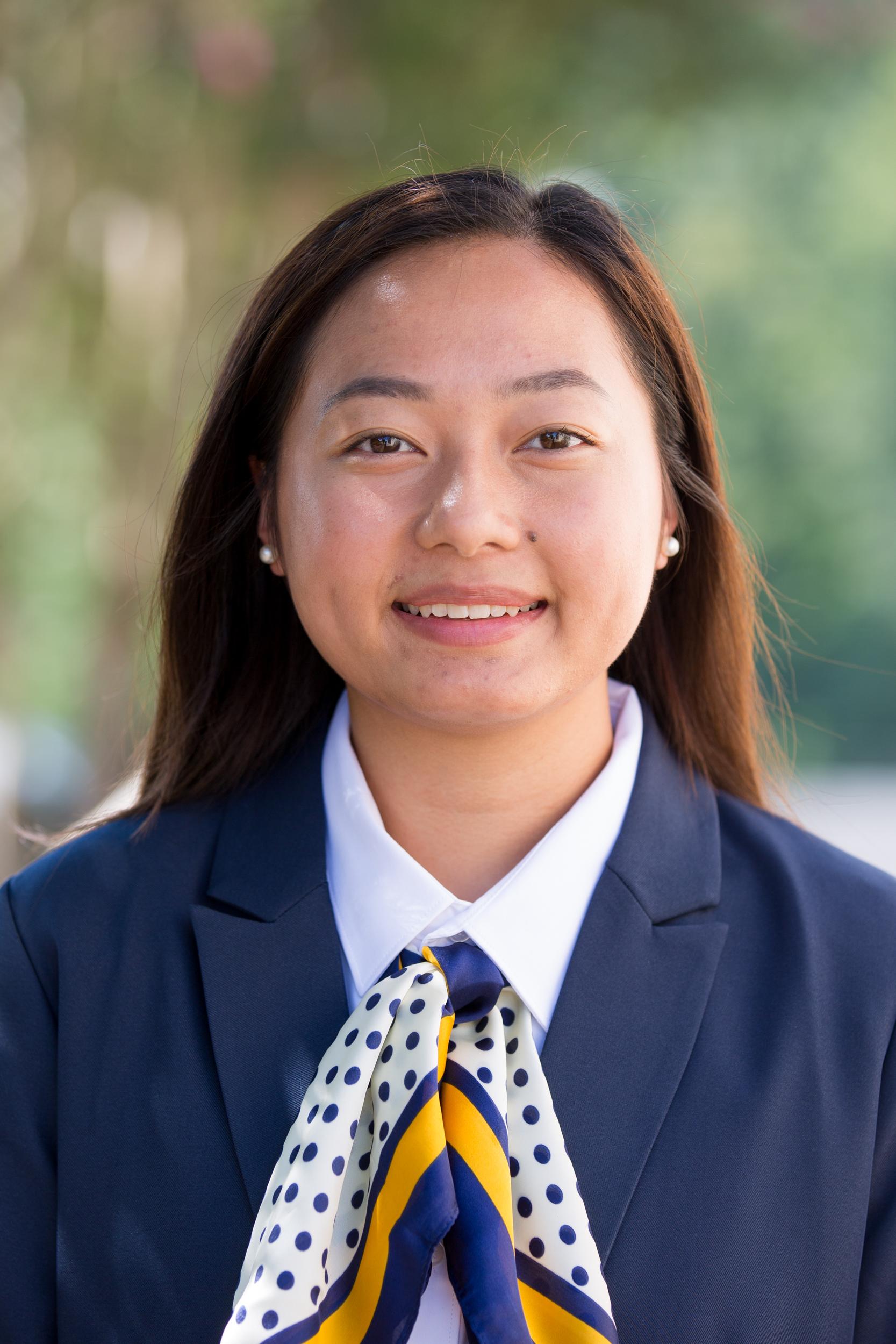 Gaoxung Lor with straight, dark brown hair, wearing a navy suit jacket, white collared shirt, a navy polka-dotted scarf, and pearl studs earrings with a small smile