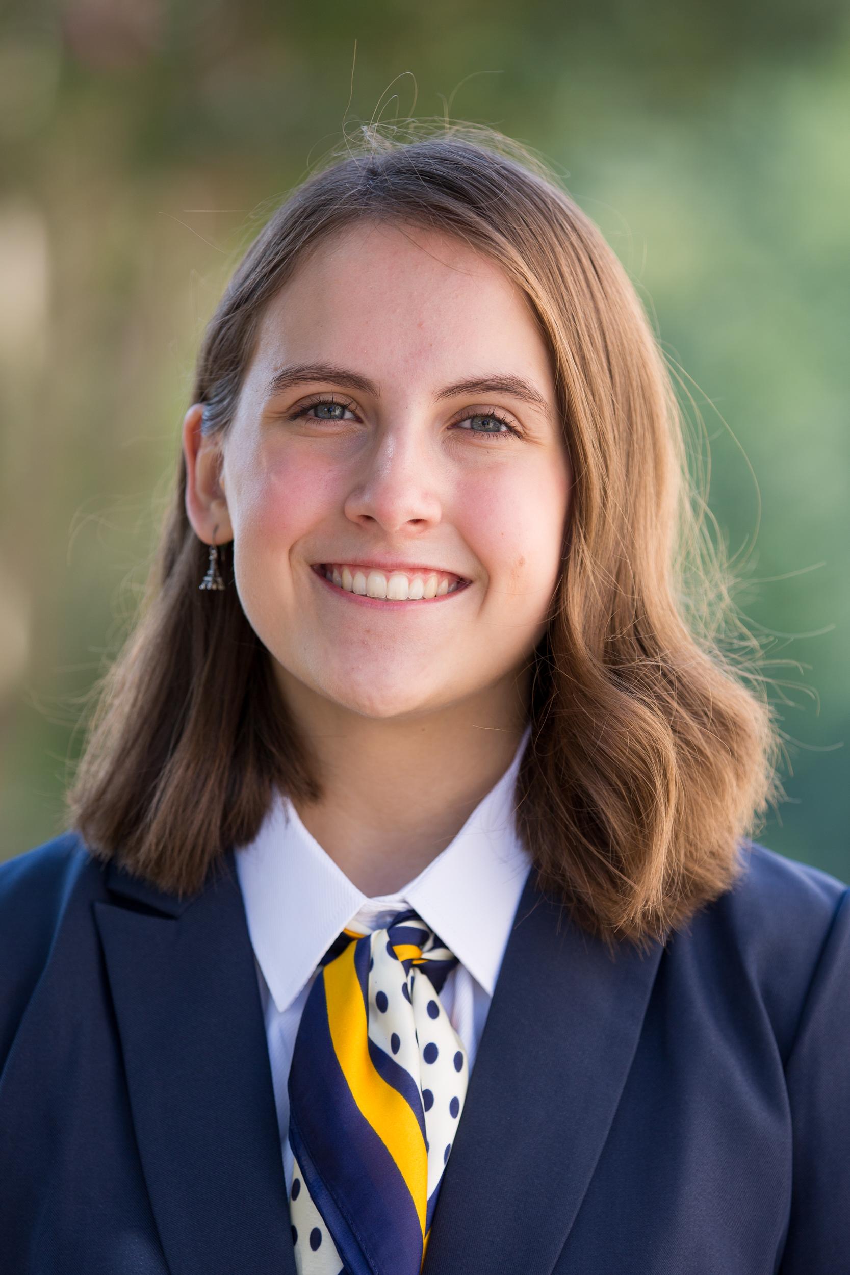 Claire Rheinecker with wavy, shoulder-length brown hair, wearing a navy suit jacket, white collared shirt, a navy, gold, and yellow scarf, and Eiffel Tower earrings, wearing a smile