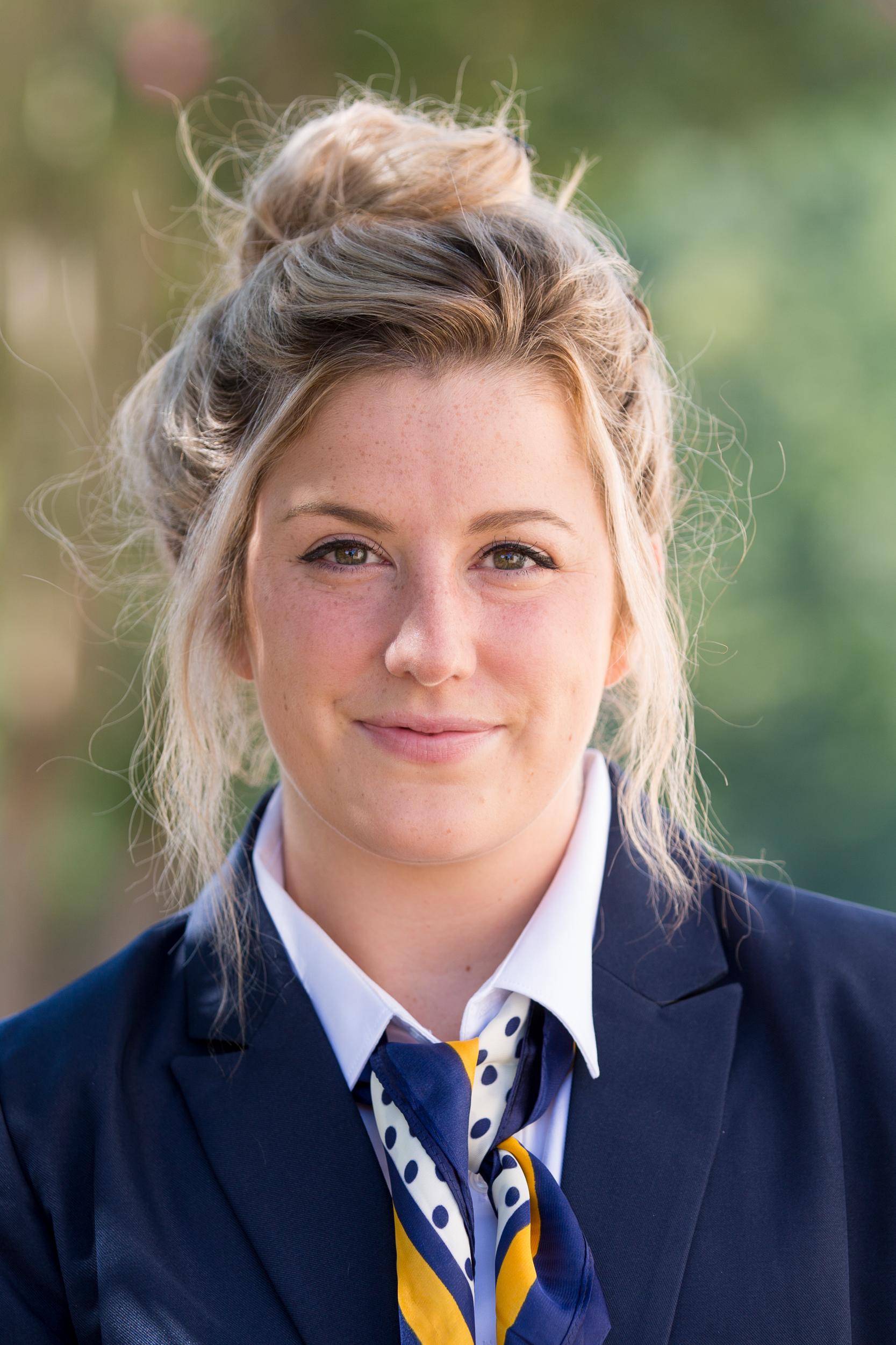 Kylie Baucom with dirty Blonde hair in a loose bun, wearing a dark blue suit jacket, white shirt, and a navy and yellow polka-dotted scarf with a closed-mouth smile