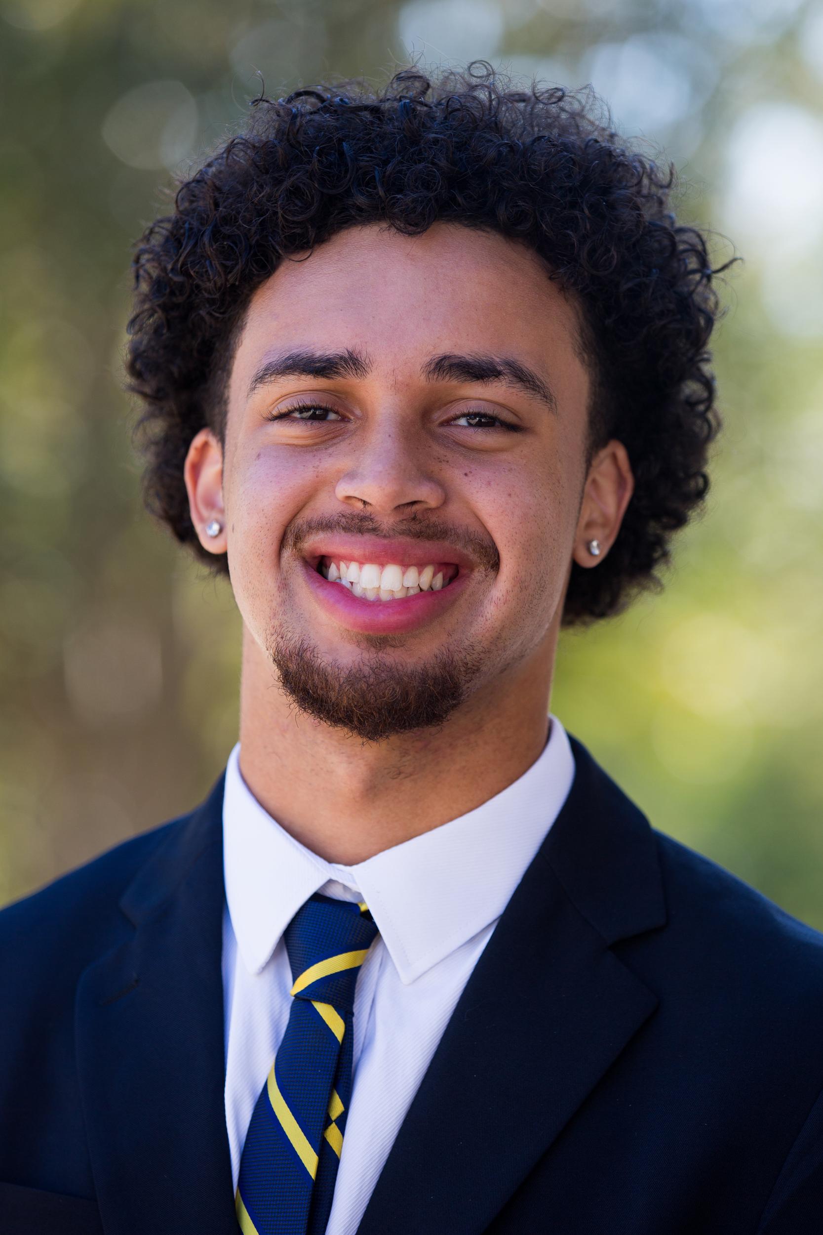 Adriel Miller with black, short curly hair and a short anchor beard, while wearing a dark navy suit and navy and gold diagonal stripe tie