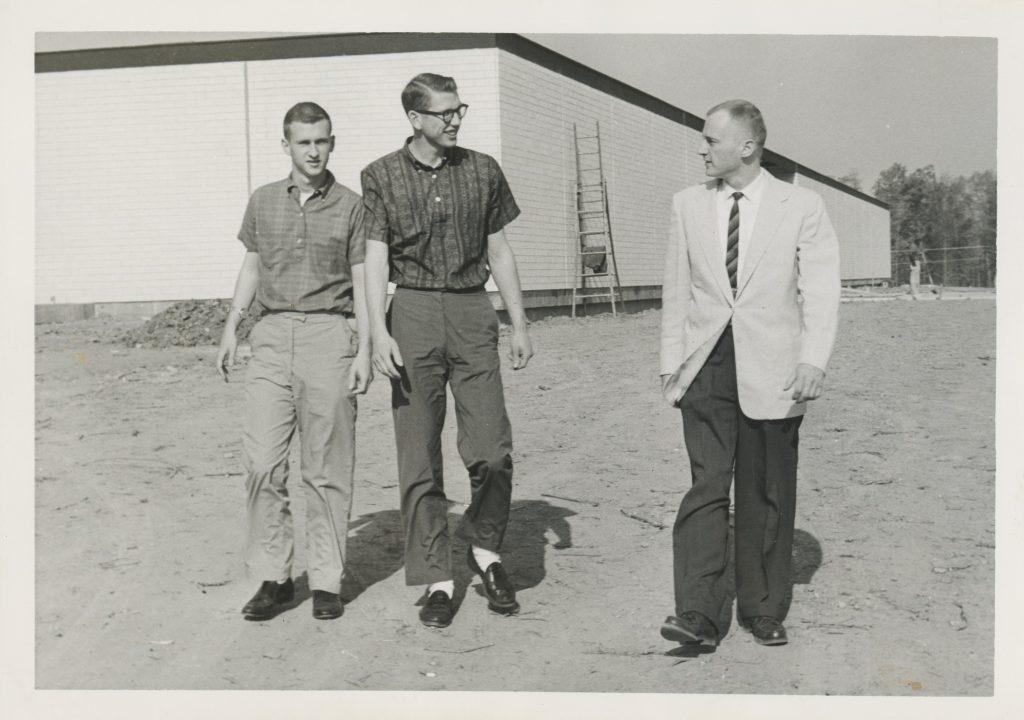 Three men walking along a dirt path beside a white building.