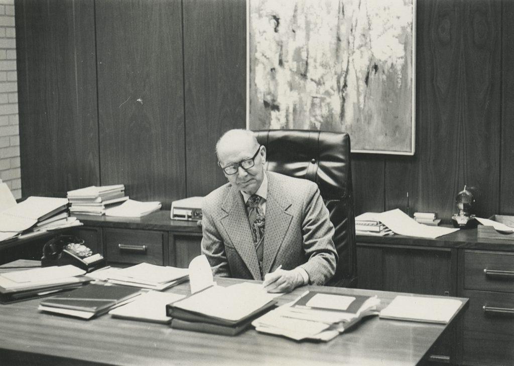 Elderly man writing at a cluttered desk in a mid-20th century office.