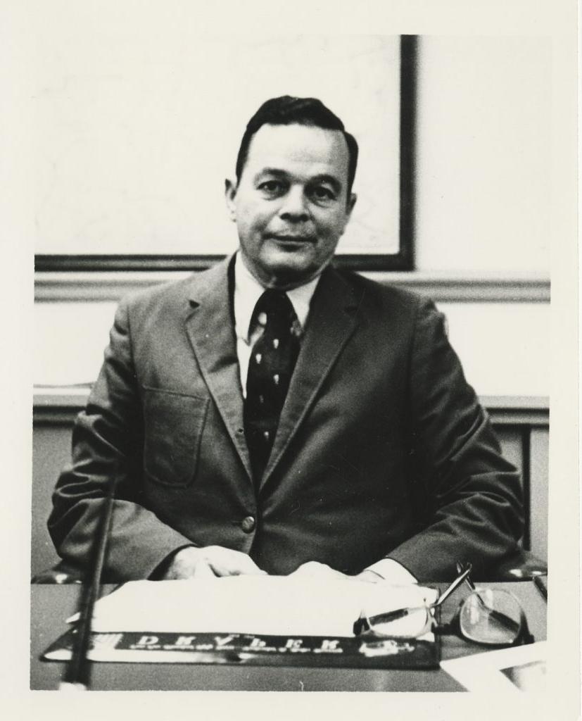 Black and white photo of William Murdoch in a suit seated at a desk with papers and glasses.