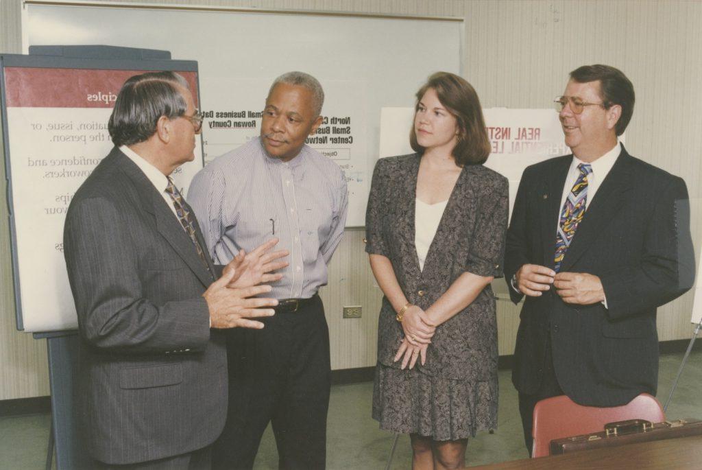 The image shows four individuals standing in a room with office furniture. Three men and one woman are engaged in conversation. The man on the far left is wearing a dark suit, white shirt, and a colorful tie, holding his hands together. Next to him stands a woman with shoulder-length brown hair, wearing a patterned blazer over a white top. She looks toward the man speaking. The third person, a man with short gray hair, wears a striped shirt and black trousers, standing with his hands in his pockets. The last man, on the right, is gesturing with his hands, wearing a dark suit with a striped tie. In the background, there's a flip chart with partially visible text and a poster board.