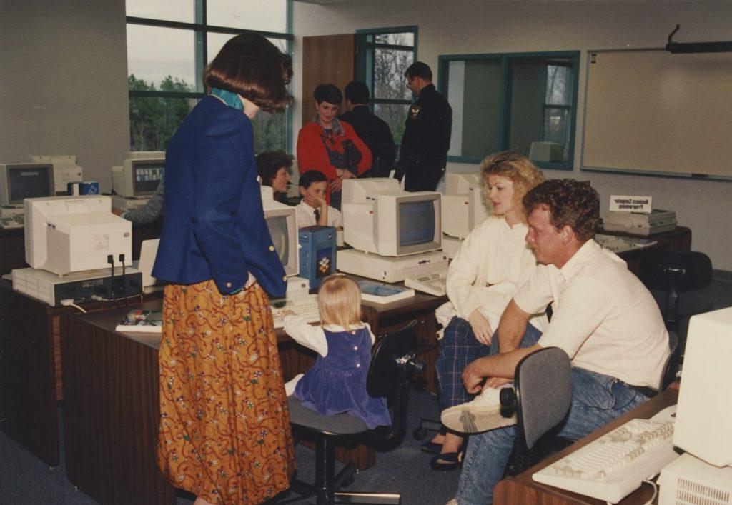 The image depicts a classroom or office setting with several people gathered around desktop computers. In the foreground, a man and woman are seated next to a young girl who is interacting with a computer. The man wears a white shirt and jeans, and the woman wears a white jacket with patterned blue pants. The girl, dressed in a purple dress, sits in a swiveling office chair, her focus directed at the monitor. Another woman, standing nearby, wears a blue blazer over a colorful patterned skirt. In the background, a group of people are engaged in conversation; one is wearing a uniform, likely a police officer. Multiple computers and printers are arranged on wooden desks throughout the room, with various office equipment visible, emphasizing a technological environment. A large window reveals a view of trees outside.