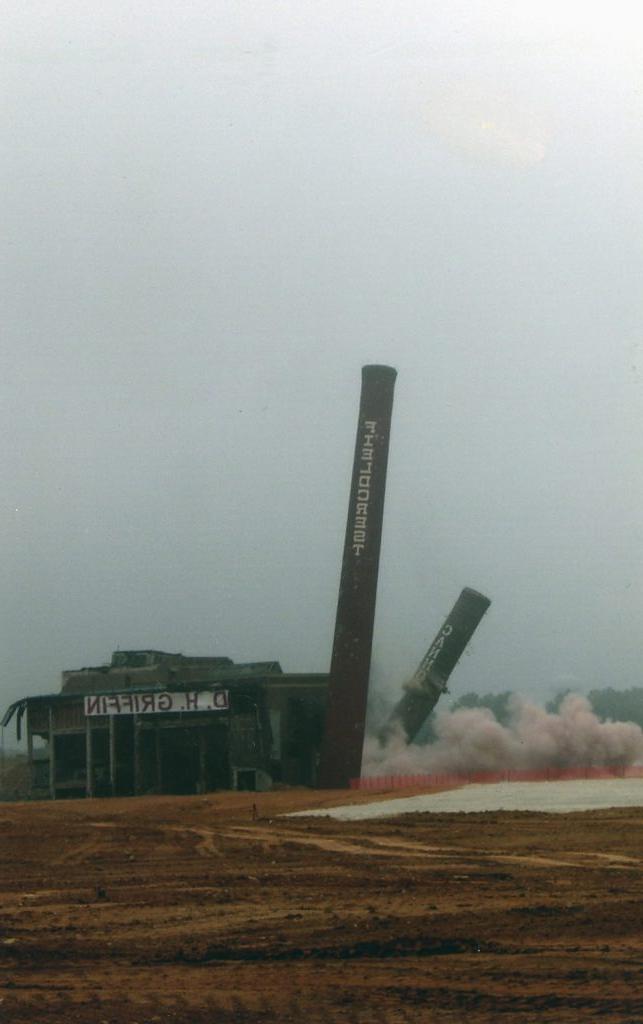 The image shows the demolition of two large industrial chimneys in a vast, open field. The main chimney, prominently featured in the center, is in mid-collapse, leaning toward the left. The second chimney is further along in its fall, closer to the ground with a cloud of dust and debris rising around its base. To the right of the image, a partially demolished industrial building is visible, with portions of the structure still standing. The sign "D. H. Griffin" is affixed to the building. The ground is barren and uneven, with reddish-brown soil visible. The sky overhead is gray and overcast, suggesting cloudy weather.